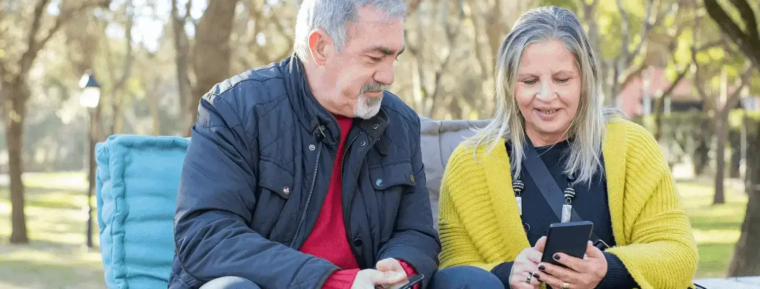two senior citizens in a park looking at their phones.