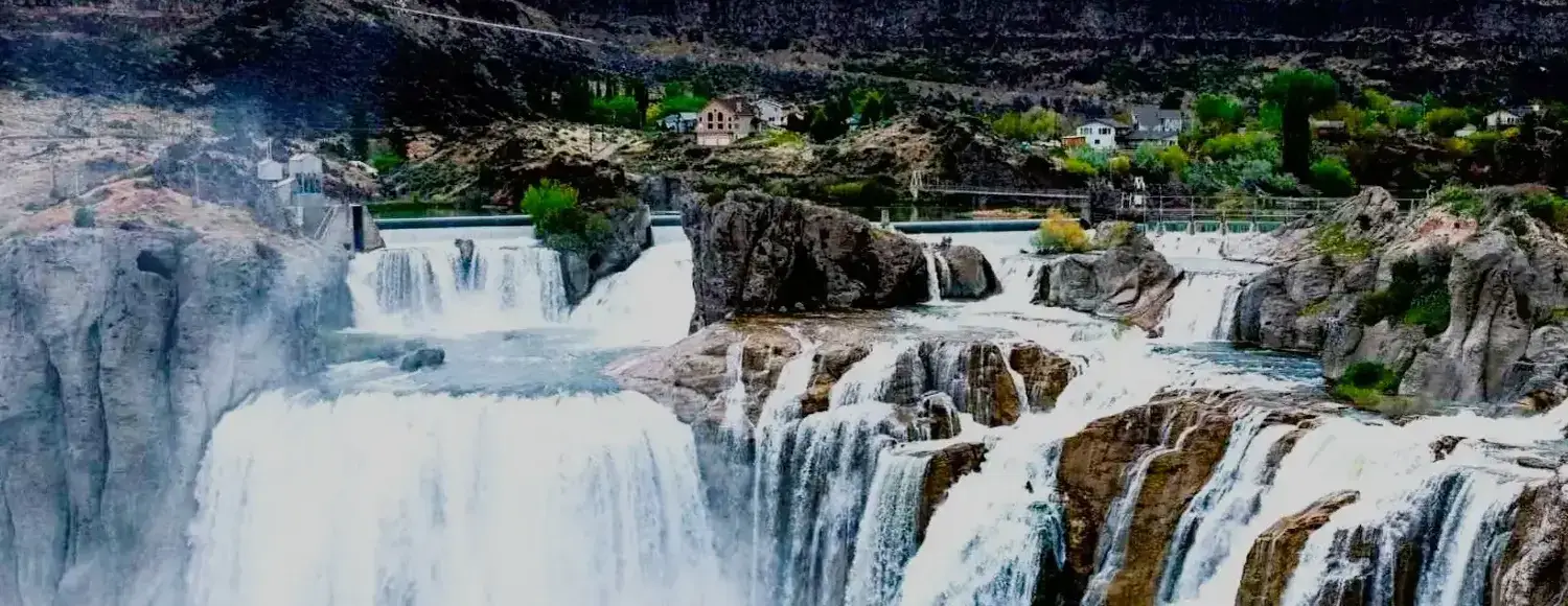 Shoshone Falls, a powerful waterfall cascading down a rocky cliff into a churning pool, surrounded by trees and a walking path. This is one of the most popular tourist destinations in Idaho.