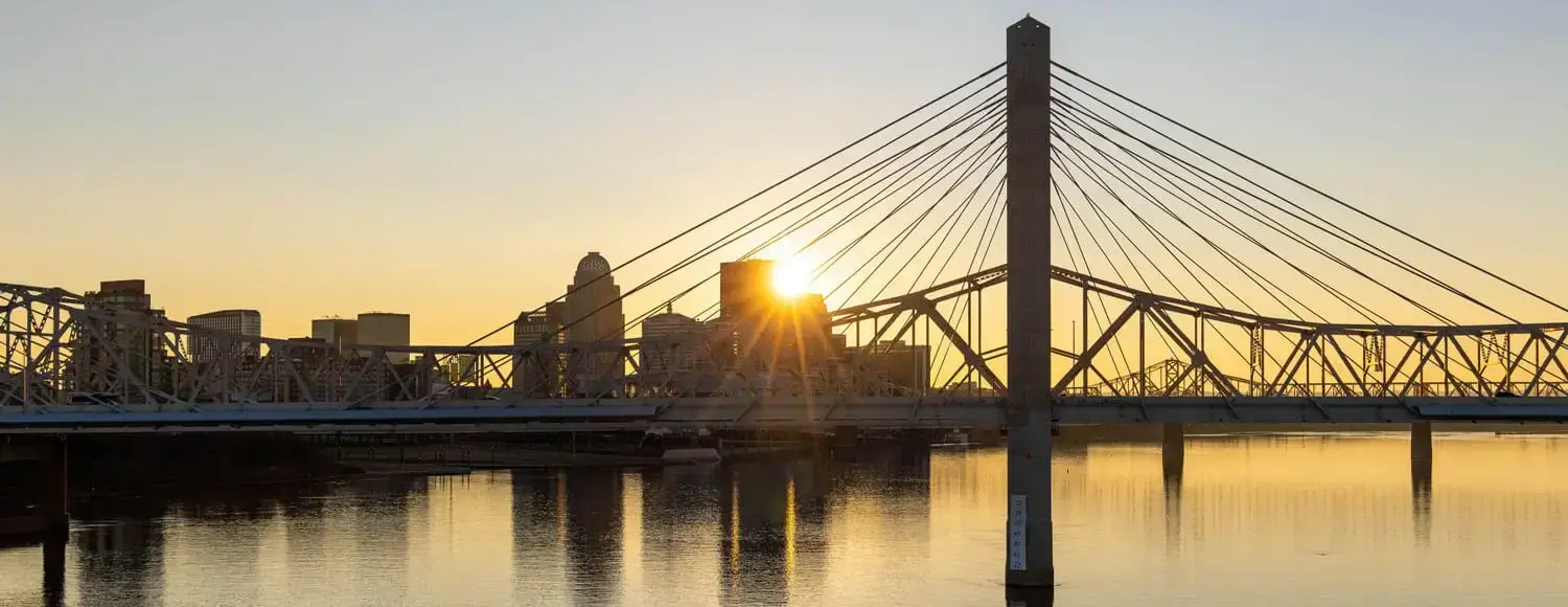 A wrought-iron truss bridge spanning the Ohio River at sunset, lit up with yellow lights. This is the Big Four Bridge in Louisville, Kentucky.