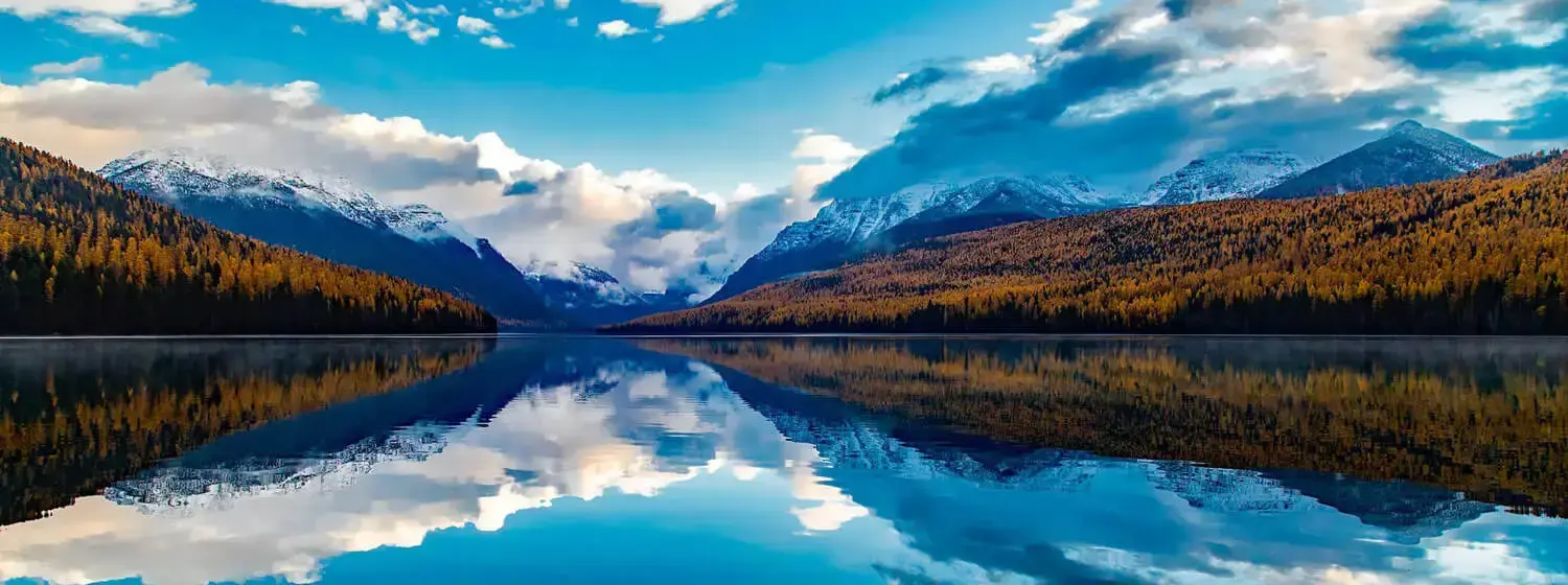 A scenic view of a lake in Montana with snow-capped mountains in the distance. The foreground shows trees lining the shore of the lake.