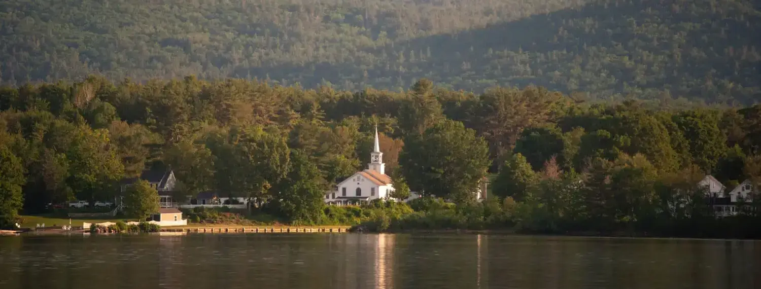 A quaint, white church with a steeple stands amongst a cluster of houses nestled within a lush green fores.