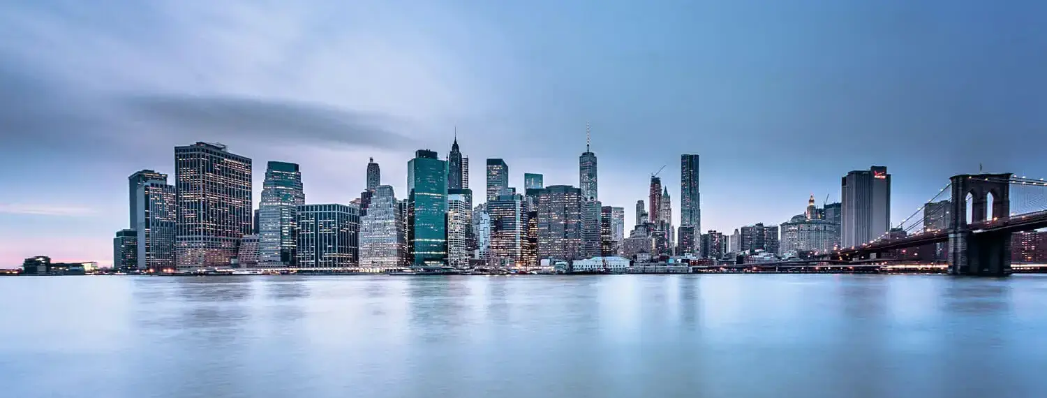 A panoramic view of New York City at sunrise, featuring the iconic Brooklyn Bridge in the foreground.