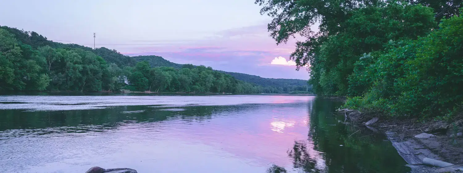 The Delaware River at sunset in Pennsylvania, with colorful rocks lining the shore and silhouettes of trees in the background.