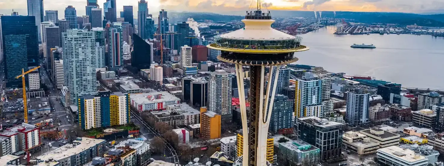 Aerial photograph of downtown Seattle showcasing skyscrapers like the Columbia Center and the Space Needle, with the waterfront of Elliott Bay and the Olympic Mountains in the background.